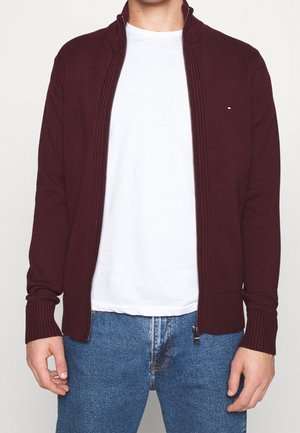 Mand iført en vinrød strikket cardigan med lynlås over en hvid t-shirt og blå jeans, stående mod en ensfarvet baggrund.