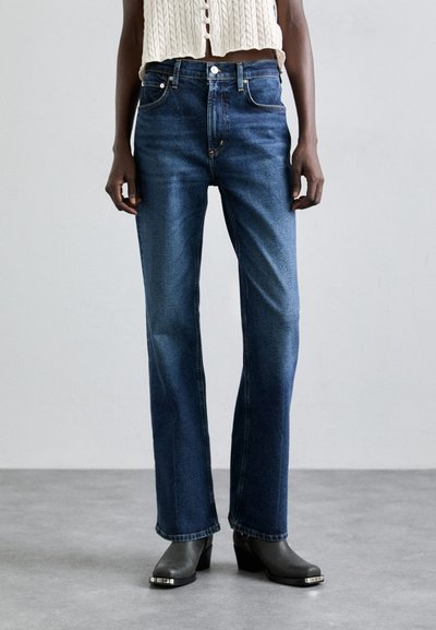 Jean en denim bleu avec une coupe droite, style à cinq poches classique et fermeture par bouton. Porté avec un top en tricot beige coupé et des bottes à talons.