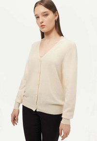 V-ringad krämfärgad cardigan i mjukt tyg, med knappstängning framtill, långa ärmar och ribbade muddar och nederkant. Bärs över mörka byxor.