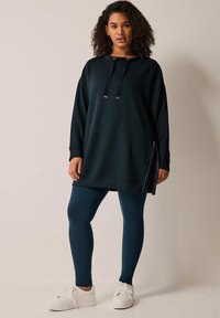 Sudadera de gran tamaño en color azul petróleo oscuro con capucha, cordones y cremalleras laterales, combinada con leggings a juego y zapatillas blancas. Textura suave.