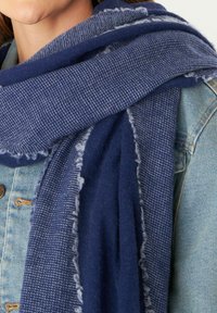 Écharpe bleu foncé présentant un motif tricot�é texturé avec des bords effilochés, superposée à une veste en denim clair. Le matériau semble doux et chaud.