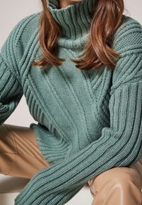 Sweter z golfem wykonany na drutach w stonowanym zielonym kolorze, z uniesionymi prążkami i grubą fakturą. Długie rękawy, luźny krój.