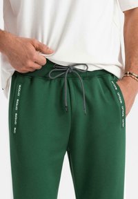 Grüne Sweatpants mit einem Kordelzugbund, ausgestattet mit Seitenpaneelen, die den Markennamen in Weiß tragen. Glatte Stoffstruktur mit einer entspannten Passform.