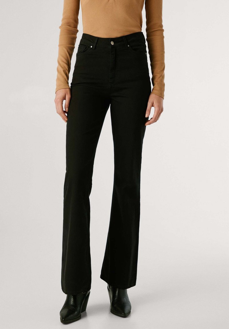 Femme portant un pantalon noir évasé taille haute, un haut à manches longues beige et des bottines à talons noires, debout devant un fond uni.