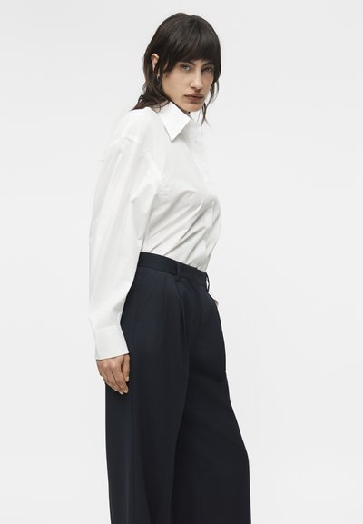 Γυναίκα με σκούρα μαλλιά, που φοράει λευκό πουκάμισο με μακριά μανίκια και μαύρο, ψηλόμεσο παντελόνι φαρδιάς γραμμής, στέκεται μπροστά σε απλό φόντο.