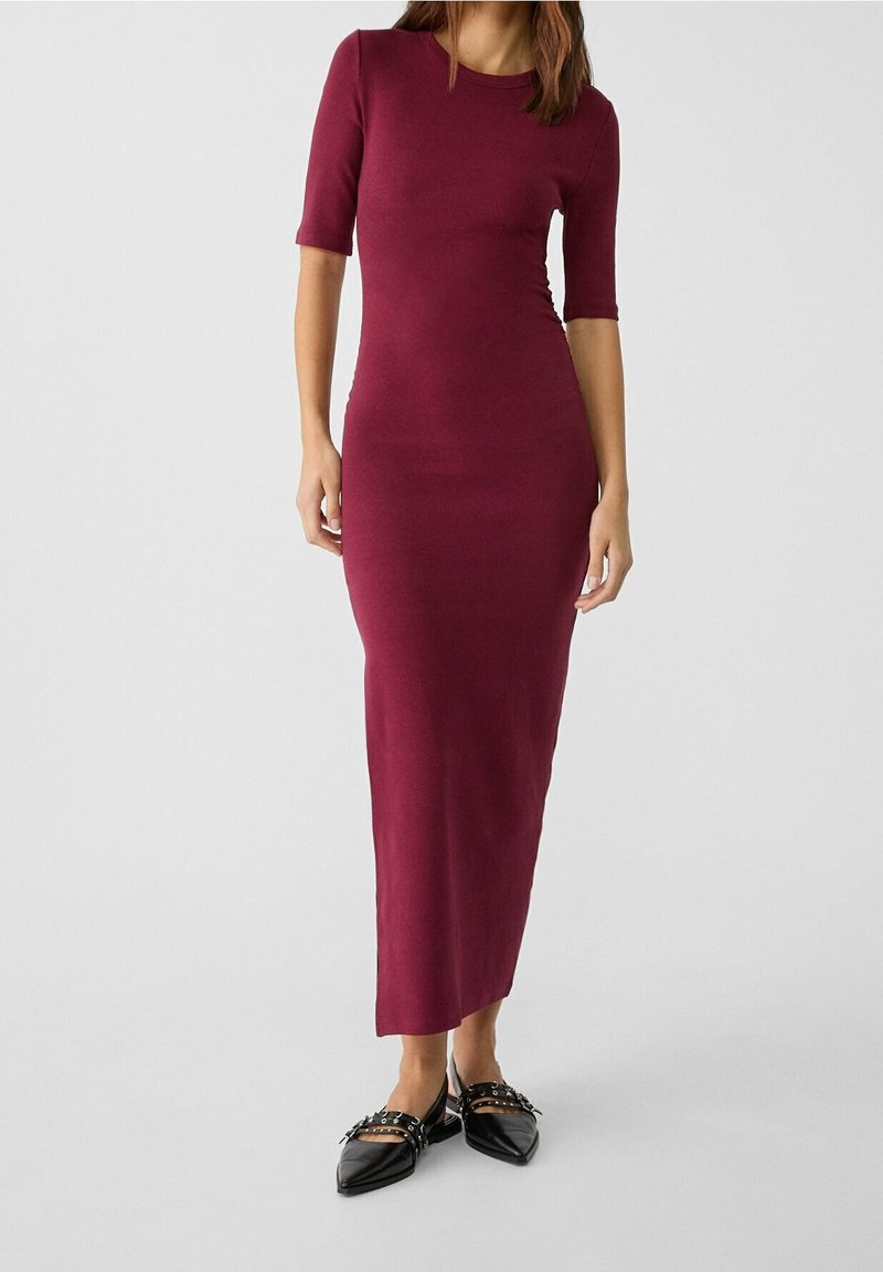 Femme portant une robe midi bordeaux ajustée avec des manches aux coudes et des ballerines noires pointues avec des brides cloutées.