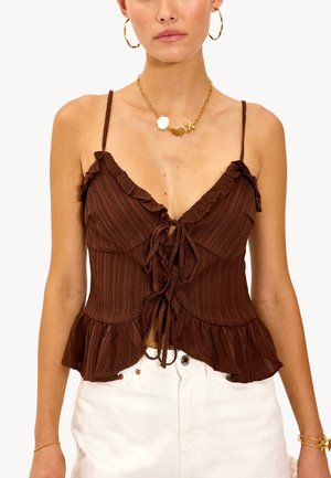 Frau mit einem braunen, rüschigen, ärmellosen Oberteil, das vorne gebunden ist, weißen, hoch taillierten Hosen, goldenen Creolen und einer goldenen Halskette mit seeinspirierten Charms.