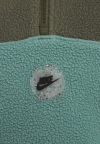 Fleece-pullover med dragkedja, med en övre del i olivgrön och en nedre del i texturerad turkos, accentuerad av en rund, prickig Nike-logotyp.