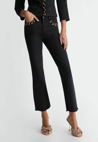 Pantalons en denim noir évasés avec un ourlet effiloché, affichant des détails en métal doré sur les poches. Portés avec des sandales à talons beige avec une lanière entrecroisée.