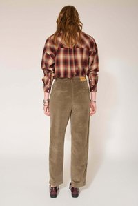 Chemise à manches longues à carreaux rouges, beige et marron, associée à un pantalon en velours côtelé vert olive avec des poches arrière et une étiquette beige. Chaussures marron.