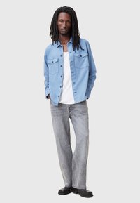 Lichtblauw overhemd met knopen en twee borstzakken, gedragen over een witte tanktop, gecombineerd met vervaagde grijze jeans en zwarte schoenen.