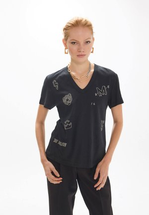 Mujer con el cabello recogido que lleva una camiseta negra de cuello en V con pequeños diseños gráficos blancos y pantalones negros, adornada con un collar y pendientes dorados.