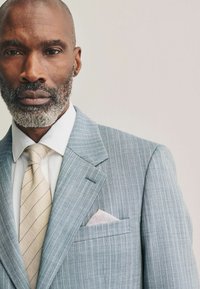 Veste de costume à rayures bleu clair avec un mouchoir de poche gris, chemise blanche et cravate à rayures beiges, mettant en valeur une coupe ajustée et un tissu texturé.