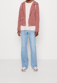 Hellrosa Reißverschlussjacke mit Kapuze, zwei Fronttaschen und Kordelzügen. Kombiniert mit einem leichten Pullover und blauen Schlagjeans. Reflektierende Turnschuhe.