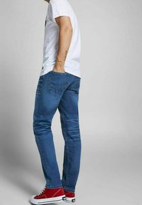 Osoba v těsných modrých džínách, bílém tričku a červených teniskách, stojící s jednou rukou v zadní kapse na jednobarevném šedém pozadí.