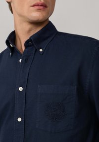 Camisa de botones azul marino, de tela de algodón, con un bolsillo en el pecho que presenta un logo bordado y detalles de textura sutil.