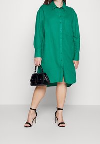 Grön button-up klänning med lång ärm, med en ficka; matchad med en svart strukturerad handväska och svarta högklackade sandaler med remmar.