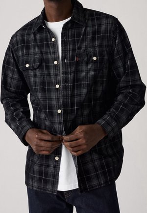 Mand, der knappet en sort og grå ternet langærmet skjorte over en hvid t-shirt, iført mørke jeans, vist fra nakke til talje.