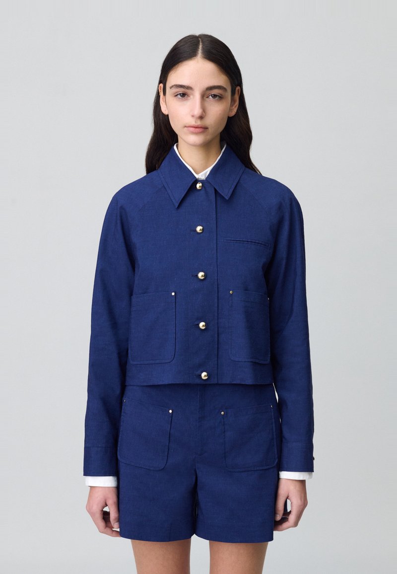 Veste courte bleu marine en tissu texturé, avec un col, des boutons dorés et deux poches avant. Portée avec un short assorti.