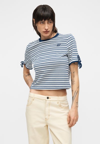 Νεαρή γυναίκα με σκούρα μαλλιά που φοράει ένα κοντό ριγέ τοπ μπλε και άσπρο και ανοιχτόχρωμο μπεζ παντελόνι, δείχνοντας τα τατουάζ στα χέρια της.