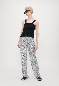 Black peplum top with adjustable straps paired with white pants featuring a black abstract pattern. Model wears black sunglasses and sandals.