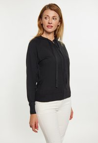 Černá mikina na zip vyrobená z lehkého materiálu, s dlouhými rukávy a šňůrkami. Má přiléhavý střih a žebrované manžety. Zobrazují se bílé kalhoty.