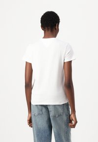 T-shirt en coton blanc à manches courtes, col rond et silhouette ajustée. Le dos présente un détail de empiècement triangulaire subtil.