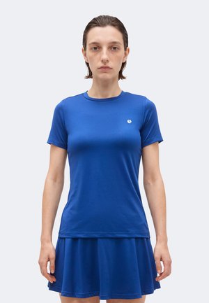 Γυναίκα με κοντά καστανά μαλλιά φοράει εφαρμοστό μπλε κοντομάνικο πουκάμισο και ταιριαστή μπλε φούστα, στέκεται μπροστά σε απλό ανοιχτό φόντο.