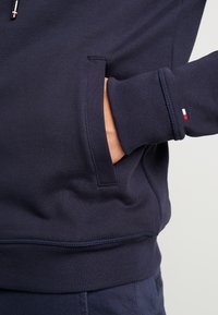 Felpa navy con tasca laterale e polsini a coste. Presenta un tessuto testurizzato con cuciture a contrasto e un piccolo logo tri-colore sulla manica.