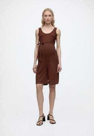 Femme portant une combinaison marron sans manches, longueur genoux, avec un lien latéral, debout devant un fond blanc uni, chaussée de sandales noires à talons.