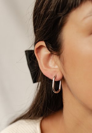Silberne Creolen mit einer länglich-oval Form und einem glatten metallischen Finish. Die Ohrringe werden am durchstochenen Ohr getragen.