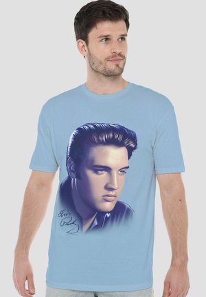 Young man wearing light blue t-shirt with large detailed portrait of a man with slicked-back hair and light blue eyes on front.