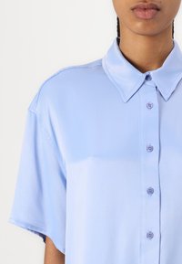 Light blue short-sleeve shirt with a classic collar, front buttons, and smooth fabric texture. Features subtle stitching details.