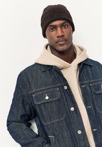 Giacca di jeans con righe verticali, due tasche sul petto, bottoni argentati. Indossata sopra una felpa con cappuccio beige e completata da un berretto a maglia marrone scuro.