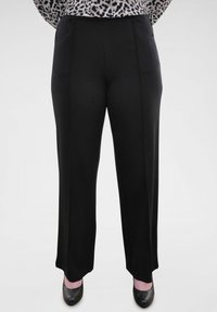 Zwarte, wijde broek van gladde stof, met twee voorzakken en een platte taille, gedragen met zwarte hakken.