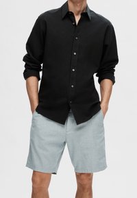 Camicia nera a maniche lunghe con le maniche arrotolate, abbinata a pantaloni corti blu chiaro. La camicia presenta un colletto e bottoni sottili di colore contrastante.