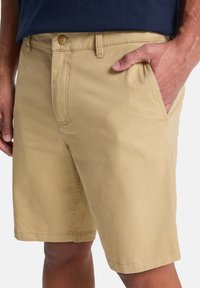 Pantaloni corti in cotone beige con chiusura a bottone, tasche laterali e design a gamba dritta, con cuciture a vista e una texture liscia.