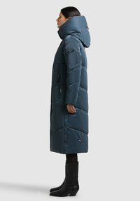 Dlouhý nadýchaný kabát tmavě modré barvy s vysokým límcem a kapucí. Má kapsu na zip a zapínání na cvočky vpředu.
