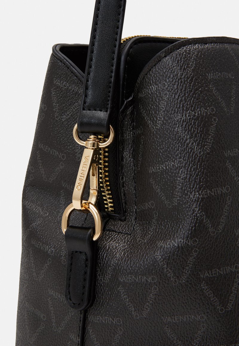 Bolso de mano de cuero negro con un sutil patrón de logo, herrajes en tono dorado, cierre de cremallera y un asa de hombro desmontable.