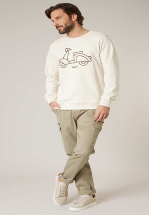 Mann trägt cremefarbenes Sweatshirt mit Rollermotiv, beige Cargohose und beige Turnschuhe, steht mit den Händen in den Taschen.