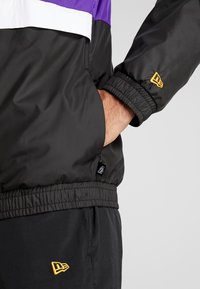 Svart och lila vindjacka med vit rand, elastiska ärm- och midjeband, med en gul broderad logotyp och fickdetaljer.