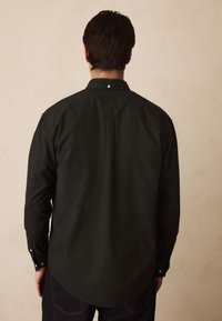 Čierna košeľa s dlhým rukávom, ktorá má špicatý golier, gombíkové manžety a detail odvetrania na chrbte. Materiál má štruktúru a pohodlný voľný strih.