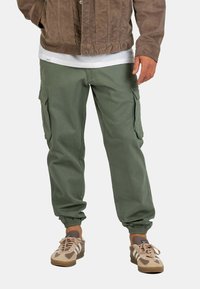 Pantalones cargo de algodón en color verde oliva, con pierna ajustada, puños elásticos y bolsillos laterales. Combinados con zapatillas deportivas en tonos beige y marrón.