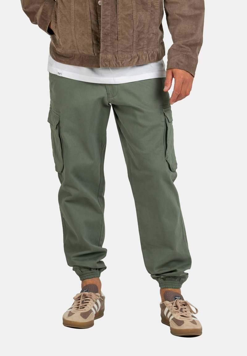 Pantalones cargo de algodón en color verde oliva, con pierna ajustada, puños elásticos y bolsillos laterales. Combinados con zapatillas deportivas en tonos beige y marrón.