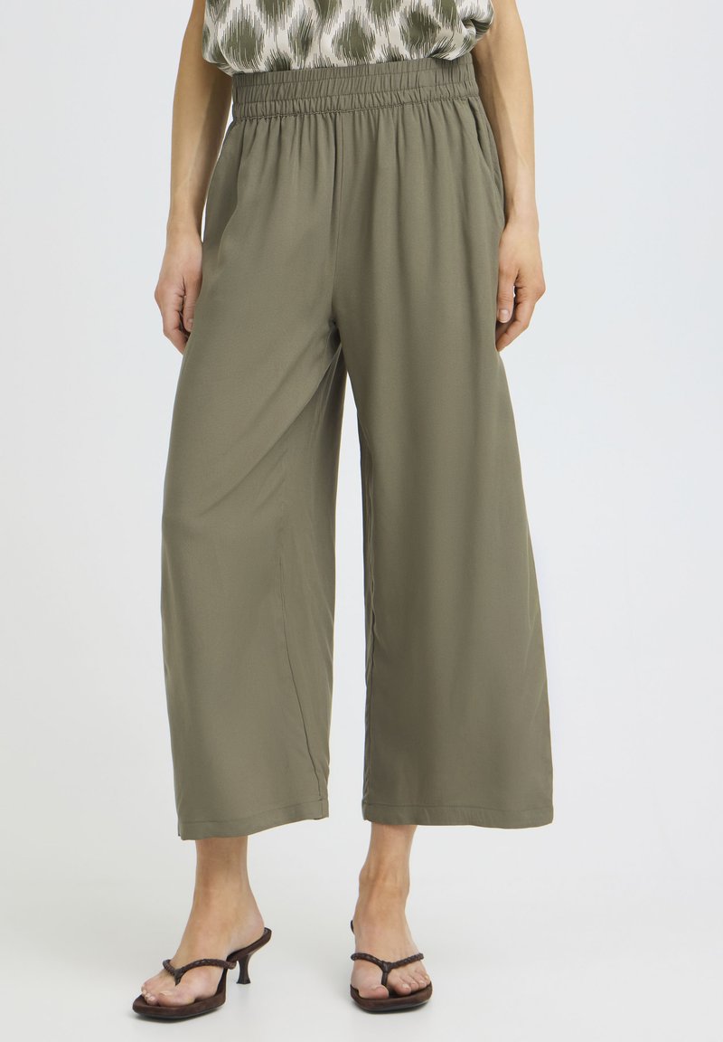 Olivrohelised laia säärega püksid, millel on elastne vöökoht, kantud beežika mustriga pealse ja pruuni kontsaga sandaalidega valgel taustal.