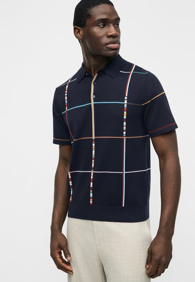 Homme portant un polo bleu marine à manches courtes avec des lignes de grille multicolores, associé à un pantalon beige clair, regardant sur le côté.