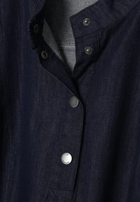 Tumši zila džinsa jaka ar daļēji atvērtām sudraba spiedpogām, kas atklāj gaišāku iekšējo audumu.