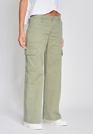 Femme portant un pantalon cargo ample vert clair avec poches à rabat et des baskets blanches, debout devant un fond uni.