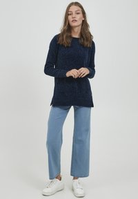Pulover tricotat bleu marin cu tivuri rigate, mâneci lungi și fante laterale, asortat cu blugi largi de culoare bleu deschis și teniși albi.