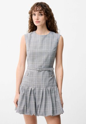 Jeune femme aux cheveux bouclés portant une robe sans manches grise à carreaux avec ceinture et jupe plissée sur fond blanc.